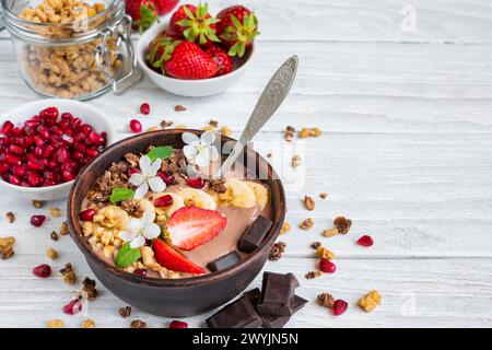 Schokoladen-Smoothie-Schüssel mit Hafergranola, Banane, Erdbeeren, Granatapfel und Frühlingsblumen auf weißem Holztisch. Gesundes veganes Protein Stockfoto