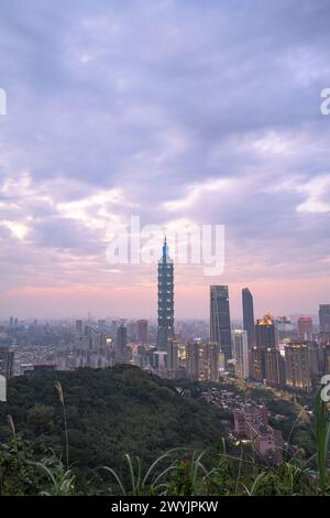 Ein erhöhter Blick auf Taipeh Stadt während der Dämmerung mit Wolkenkratzern vor einem subtilen Sonnenuntergang, aufgenommen von einem bergigen Gebiet mit Laub Stockfoto