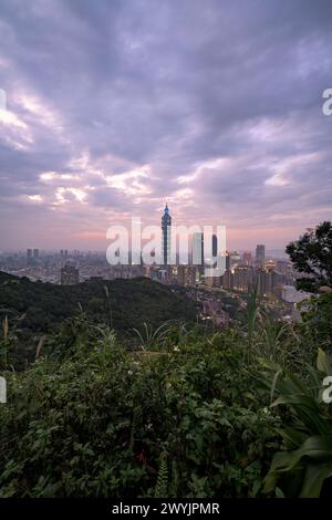 Ein erhöhter Blick auf Taipeh Stadt während der Dämmerung mit Wolkenkratzern vor einem subtilen Sonnenuntergang, aufgenommen von einem bergigen Gebiet mit Laub Stockfoto