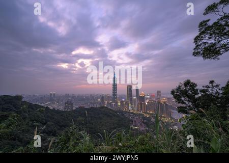 Ein erhöhter Blick auf Taipeh Stadt während der Dämmerung mit Wolkenkratzern vor einem subtilen Sonnenuntergang, aufgenommen von einem bergigen Gebiet mit Laub Stockfoto