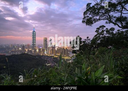 Ein erhöhter Blick auf Taipeh Stadt während der Dämmerung mit Wolkenkratzern vor einem subtilen Sonnenuntergang, aufgenommen von einem bergigen Gebiet mit Laub Stockfoto