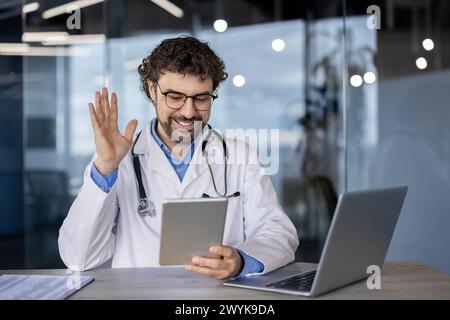 Lächelnder männlicher Arzt im Laborkittel, der während einer telemedizinischen Konsultation winkt, die Zugänglichkeit und moderne Gesundheitsdienste zum Ausdruck bringt. Stockfoto