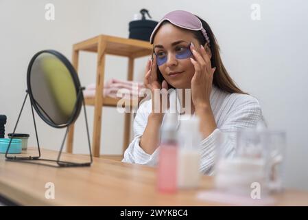 Die Dame in einem Bademantel trägt Augenflecken und Hautpflege-Routine auf, während sie während eines Spa-Tages in den Spiegel blickt. Stockfoto