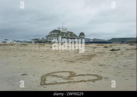 2023 Hampton Beach, New Hampshire. Ein großes Herz ist in den Sand gezeichnet, in dem die Nachricht Help mit dem Seashell Bühnengebäude im geschrieben ist Stockfoto