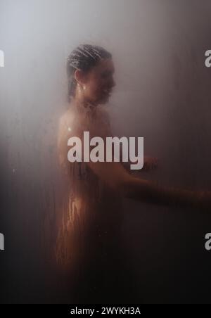 Junge Frau hinter dampfendem Glas im Badezimmer, die heißes Wasser beim Duschen anpasst. Stockfoto