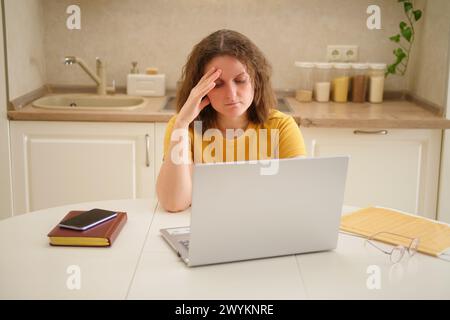 Eine traurige Frau mit Laptop sitzt an einem Tisch in einer Küche. Eine erwachsene Geschäftsfrau arbeitet von zu Hause aus, in einem entfernten Büro Stockfoto