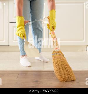 Eine Frau fegt den Boden mit einem Besen und einer Schmutzpfanne in ihrer Küche. Weibliche Hände in gelben Handschuhen beim Reinigen der Küche Stockfoto