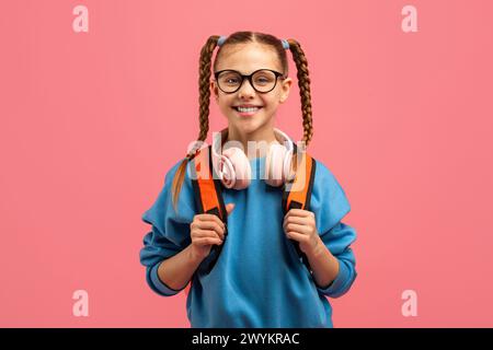 Lächelndes Mädchen mit Kopfhörern und Rucksack auf rosa Hintergrund Stockfoto