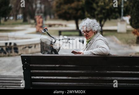 Aktive Seniorin mit Fahrrad, die mit Smartphone auf Parkbank sitzt Stockfoto