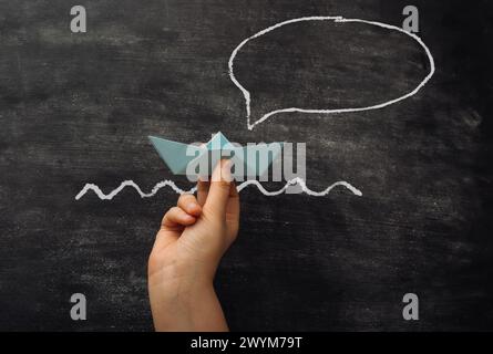 Eine Hand, die ein Papierboot vor einer Tafel mit einer Sprechblase hält. Die Sprechblase sagt, ich bin ein Boot. Stockfoto