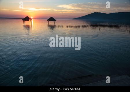 Ein wunderschöner Sonnenuntergang über dem See Peten Itza im Norden Guatemalas wirft orange rosa und blaue Farbtöne über das ruhige Wasser, mit Hütten im Wasser Stockfoto