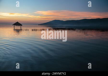 Ein wunderschöner Sonnenuntergang über dem See Peten Itza im Norden Guatemalas wirft orange rosa und blaue Farbtöne über das ruhige Wasser, mit Hütten im Wasser Stockfoto