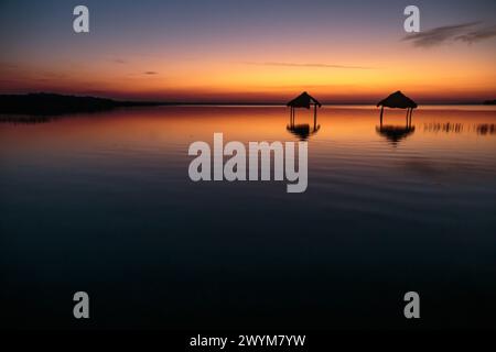 Ein wunderschöner Sonnenuntergang über dem See Peten Itza im Norden Guatemalas wirft orange rosa und blaue Farbtöne über das ruhige Wasser, mit Hütten im Wasser Stockfoto