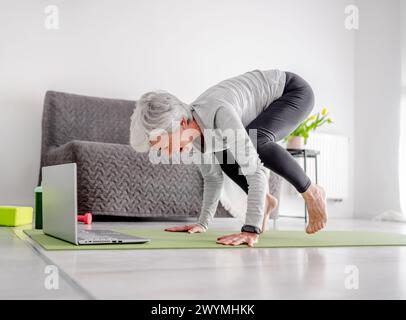 Angenehme 70-Jährige Frau Führt Komplexe Übungen Mit Trainer Über Laptop Durch Und Macht Yoga Zu Hause Online Stockfoto