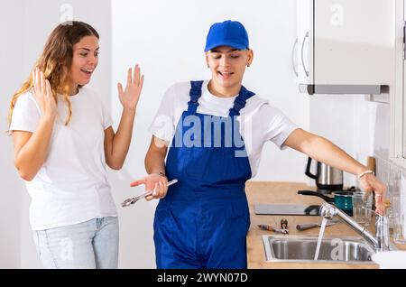 Glückliche, zufriedene Hausfrau überprüft den Betrieb des Wasserhahns in der Küche und danke dem Klempner Stockfoto