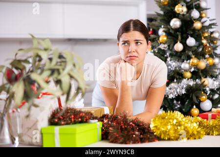 Emotionale traurige junge Frau neben einer verpackten Geschenkbox mit Schleife in der Nähe und dekoriertem Weihnachtsbaum zu Hause Stockfoto