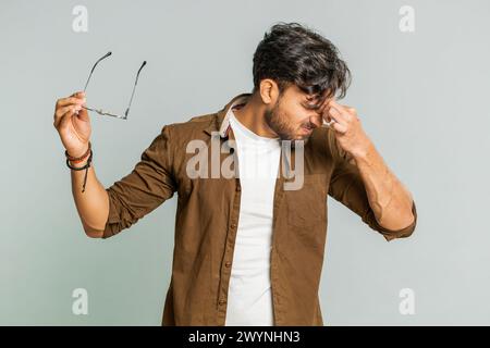 Erschöpfter, müder indischer junger Mann zieht die Brille aus, spürt Augenschmerzen, ist überlastet Burnout von langer Arbeit. Verschlafener, erschöpfter Arabianer, der die Augen isoliert auf grauem Hintergrund reibt Stockfoto