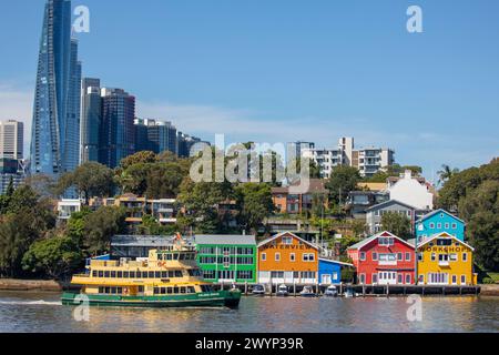 Farbenfrohe historische Waterview Wharf Werkstattgebäude an der Uferpromenade auf der Balmain Peninsula, vorbeifahrende Fähren von Sydney und Sydney Wolkenkratzer Stockfoto