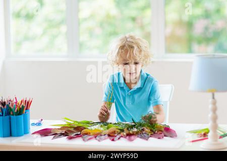 Kind schafft Bild mit bunten Blättern. Kunst und Kunsthandwerk für Kinder. Kleiner Junge, der Collagebild mit Regenbogenpflanzen-Blatt macht. Hausaufgaben für die Biologie Stockfoto