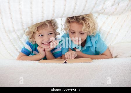 Lesebuch für Kinder im Bett unter der Strickdecke. Zwei Brüder spielen zusammen. Kinder gemütliches Zimmer im Hyggge-Stil. Kleiner Junge, der Hausaufgaben macht, bevor er schläft. Stockfoto