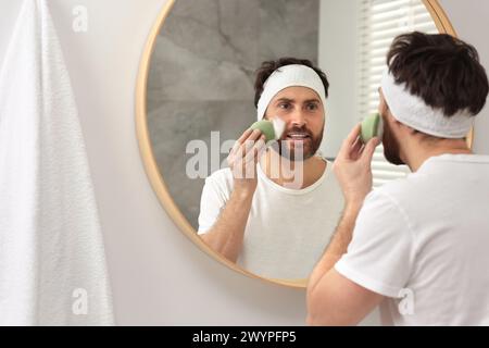 Mann mit Stirnband, der sein Gesicht mit einem Schwamm in der Nähe des Spiegels im Badezimmer wäscht Stockfoto