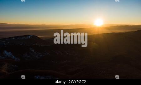 Die Sonne geht über einer ruhigen Berglandschaft auf und wirft ein warmes Leuchten über die nebeligen Täler und hügeligen Hügel, die in einem weiten Blick aus der Luft erfasst werden. Stockfoto