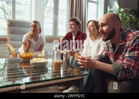 Angespannter Spielmoment. Gruppe von Jugendlichen, Freunden, die sich zu Hause treffen, im Wohnzimmer sitzen, Bier trinken und Online-Matchübersetzungen anschauen. Stockfoto