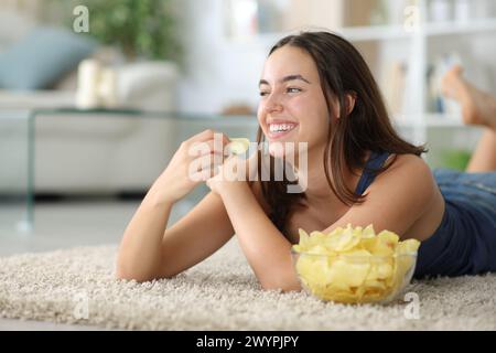 Glückliche Frau isst Kartoffelchips, die zu Hause auf einem Teppich liegen und auf die Seite schauen Stockfoto