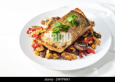 Regenforellenfilet mit knuspriger frittierter Haut auf Gemüse aus dem Ofen und Petersiliengarnitur serviert auf einem weißen Teller, gesundes Fischgericht geeignet für Low Stockfoto