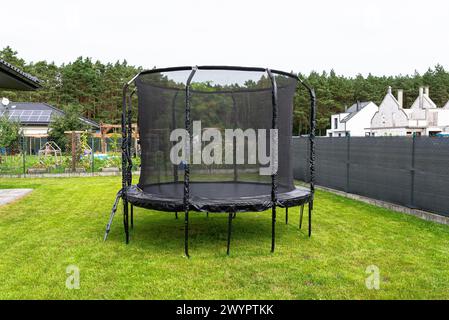 Großes Springtrampolin für Kinder mit Schutznetz und geschlossenem Reißverschluss, im Garten stehend, sichtbares Netz, das den Zaun bedeckt. Stockfoto