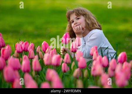 Fröhliches Kind, das im grünen Gras sitzt und sich pinkfarbene Tulpen ansieht. Niedliches Kind spielt im Frühlingsblütenfeld. Glückliches Kind, das allein im Frühlingsgarten spielt. Stockfoto