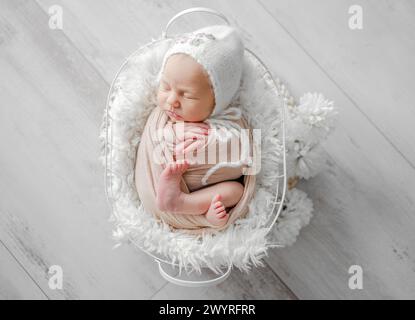 Das Neugeborene Mädchen, das in Eine Decke und einen Hut gehüllt ist, schläft in Einem beigefarbenen Korb und wird in Einem professionellen Neugeborenen-Fotoshooting festgehalten Stockfoto