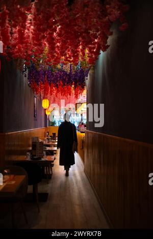 Eine junge Frau, die von hinten gesehen wird, verlässt ein japanisches asiatisches Restaurant. Sie ist im Schatten hinterleuchtet und die Blumendekoration an der Decke verleiht Farbe Stockfoto