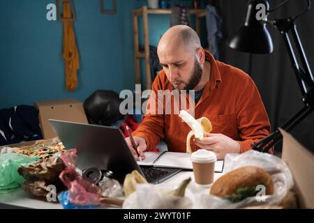 Freiberuflicher Mitarbeiter, der zu Hause in einem schmutzigen, überfüllten Raum arbeitet, Mann, der einen Laptop benutzt, isst Bananen am Arbeitsplatz Stockfoto