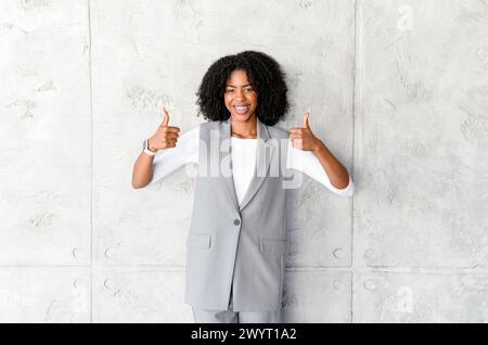 Eine bestrahlende afroamerikanische Geschäftsfrau zeigt sich doppelt hoch und signalisiert Anerkennung und Erfolg vor einem grauen Hintergrund, der Einfachheit und Konzentration vermittelt. Stockfoto