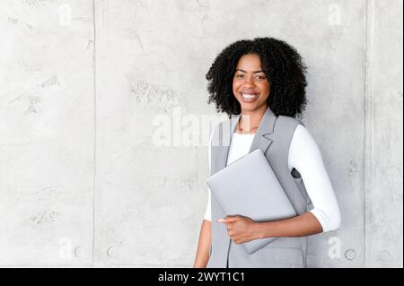 Eine geschickte afroamerikanische Geschäftsfrau steht selbstbewusst mit ihrem Laptop, ihr Lächeln strahlt Professionalität vor einem minimalistischen Hintergrund aus. Dieses Bild spiegelt das Wesen des modernen Unternehmenserfolgs wider Stockfoto