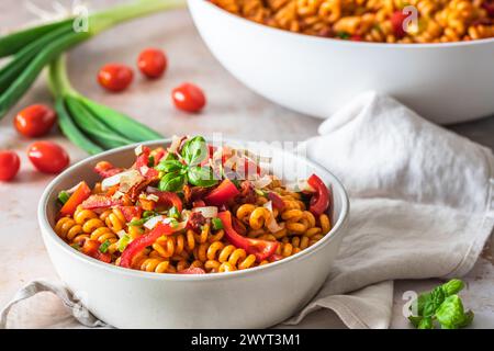 Nudelsalat mit rotem Pfeffer, Tomaten, Frühlingszwiebeln und Basilikum in einer weißen Schüssel auf hellem Hintergrund Stockfoto