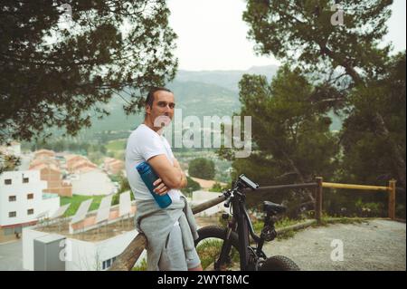 Schöner muskulöser Mann in weißem T-Shirt und grauen Shorts, der neben seinem Elektro-Mountainbike an einem Holzzaun steht, auf einer Landstraße mit Blick auf die Weiße Stockfoto