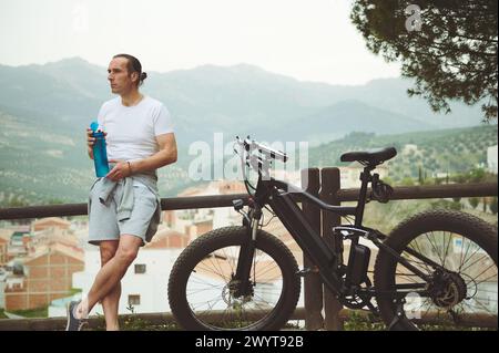 Kaukasischer Athletiker, der eine Flasche Wasser hält, träumerisch aussehend, entspannend nach dem Radfahren in den Bergen, am Holzzaun in der Nähe seines elektrischen Stroms stehend Stockfoto