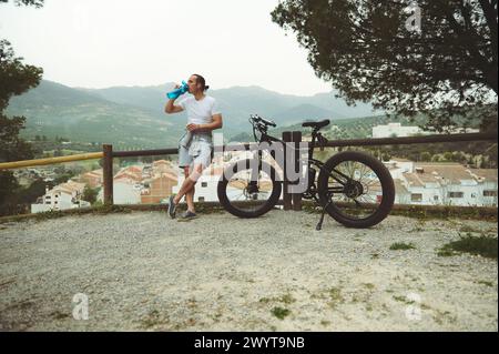 Ein gutaussehender muskulöser Radfahrer trinkt Wasser aus einer Sportflasche, steht neben einem Holzzaun neben dem E-Mountainbike auf einer Landstraße mit Blick auf wh Stockfoto