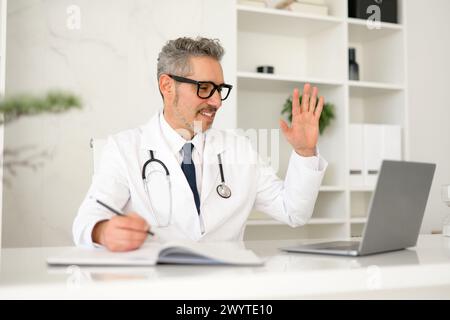 Fröhlicher Seniorarzt arbeitet virtuell mit Patienten oder Kollegen zusammen, indem er Handgesten verwendet, um während einer Online-Konsultation oder eines Meetings von seinem Büro aus zu erklären oder zu kommunizieren. Moderne Telemedizin Stockfoto