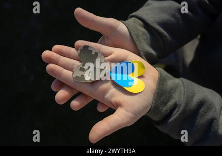 Herzausschnitt aus militärischem Tarnstoff und gelbem blauem Papier liegt auf der Kinderhandfläche. Stoppt den Krieg in der Ukraine. Mit Liebe und Dankbarkeit den Soldaten der Th Stockfoto