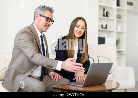 Ein leitender Geschäftsmann spricht mit einer jungen Frau, die beide Interesse an Inhalten zeigen, die im Büro auf einem Laptop angezeigt werden. Die Erfahrung trifft auf Jugendliche Energie in einem produktiven Austausch Stockfoto