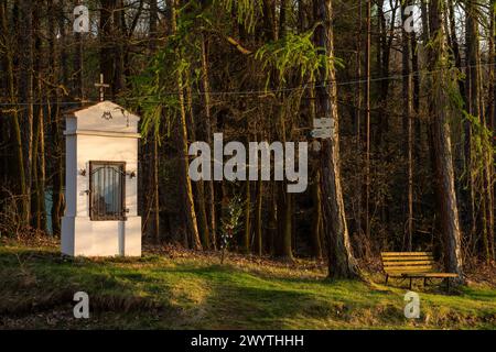 Kleine ländliche Kapelle in Zátor, Brdy Hills, Tschechien. Wald im Hintergrund, Touristenpfade Schilder hängen an einem Baum und eine Bank steht in der Nähe. Stockfoto