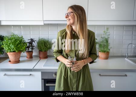 Zufriedene Geschäftsfrau in Gläsern mit Glas Wasser, die in der Küche zu Hause steht Stockfoto