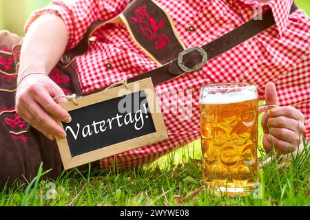 Bayern, Deutschland - 7. April 2024: Vatertag-Schriftzug auf einem Brett, gehalten von einem Mann in bayerischer Lederhose und traditionellem Hemd zusammen mit einem Becher Bier. FOTOMONTAGE *** Vatertag Schriftzug auf einer Tafel die ein Mann in bayerischer Lederhose und Trachtenhemd zusammen mit einem Maßkrug Bier hält. FOTOMONTAGE Stockfoto