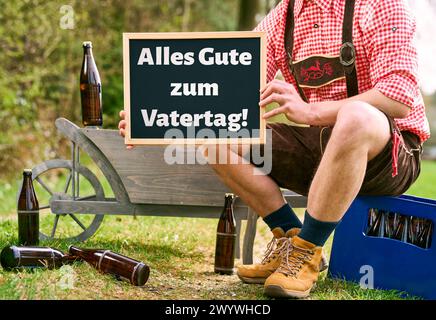 Bayern, Deutschland - 7. April 2024: Mann in Lederhosen auf einem Vatertagsausflug mit Bierkisten und Handkarren mit einem Schild mit der Aufschrift: Happy Vatertag. Grüße zum Vatertag. FOTOMONTAGE *** Mann in Lederhose bei einem Vatertagsausflug mit Bierkisten und Bollerwagen hält ein Schild mit Aufschrift: Alles Gute zum Vatertag. Vatertagsgrüße. FOTOMONTAGE Stockfoto