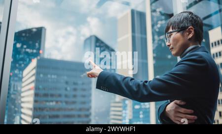Stilvoller japanischer Geschäftsmann, der mit dem Glasaufzug zum Büro im modernen Business Center in der Innenstadt fährt. Junger erfolgreicher asiatischer Mann, der mit Mock-up-Holographic Screen im Lift-Konzept interagiert. Stockfoto