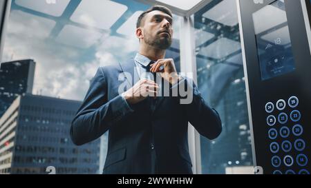 Erfolgreicher Geschäftsmann im Anzug, der Krawatte fixiert, während er mit dem Glasaufzug zum Büro im modernen Business Center fährt. Vorbereitung auf wichtige Besprechungen Stockfoto