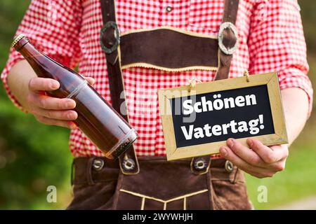 Bayern, Deutschland - 7. April 2024: Junger Mann in bayerischer Tracht hält eine Flasche Bier und ein Schild mit dem Gruß: Happy Vatertag. FOTOMONTAGE *** junger Mann in bayerischer Tracht hält eine Flasche Bier und ein Schild mit dem Gruß: Schön Vatertag. FOTOMONTAGE Stockfoto